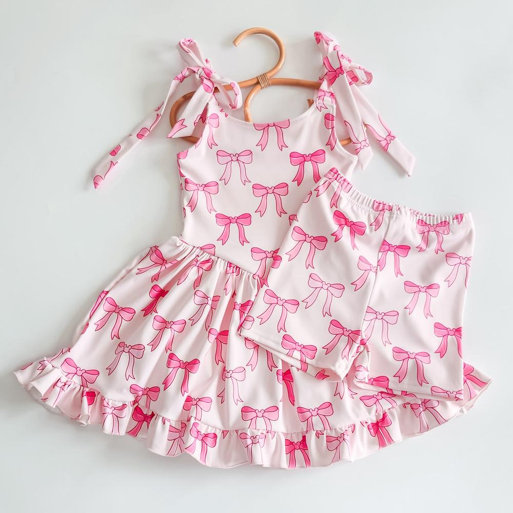 Pink activewear dress and biker shorts set with a playful bow print, ruffled hem, and shoulder ties.