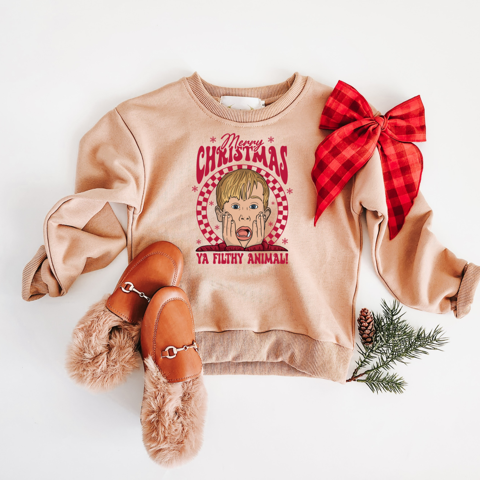 Beige sweatshirt with graphic design and red bow, paired with brown shoes and pine branches on a white background