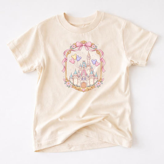 Enchanted Kids T-shirt: beige tee with whimsical park castle design, pink bow, balloons, and flowers.