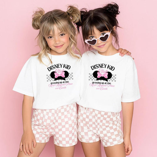 Two girls in white "The Park Kid Club" t-shirts with pink bows and checkered shorts.