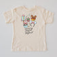 Kids' cream t-shirt with "Infinity Snacks and Beyond" text and colorful cartoon snacks like a rocket, boba, and The waffle.