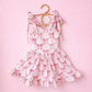 White activewear dress with pink bow print, shoulder ties, and ruffled hem, hanging on a rattan hanger.