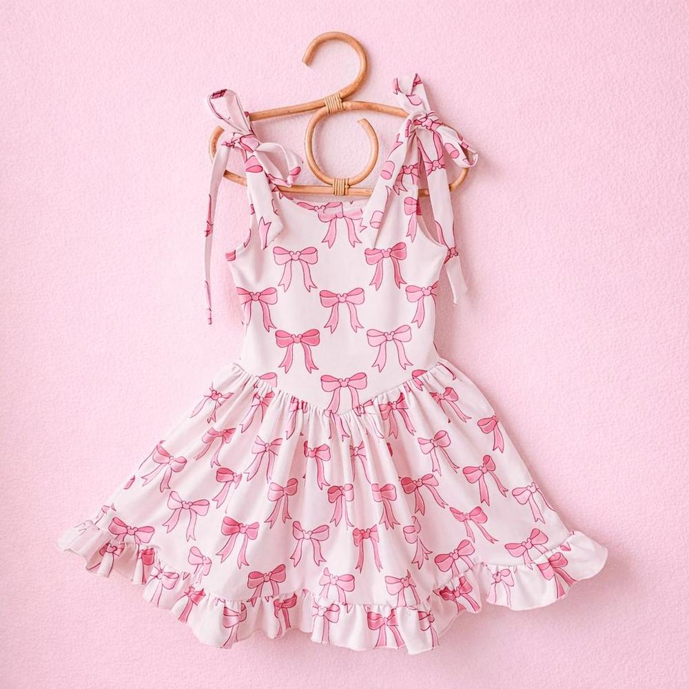 White activewear dress with pink bow print, shoulder ties, and ruffled hem, hanging on a rattan hanger.