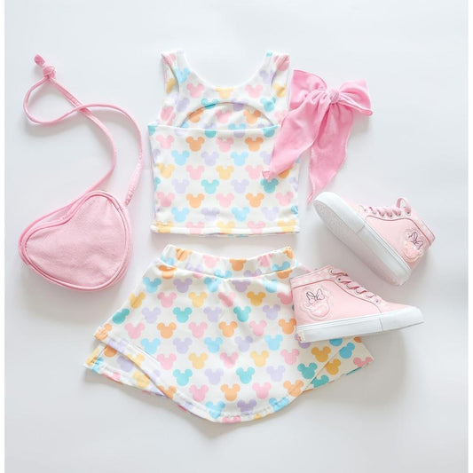 Kids' two-piece athleticwear: white crop top and skirt with attached biker shorts, featuring pastel The Mouse heads, paired with pink high-top sneakers, a heart-shaped purse, and a large pink bow.