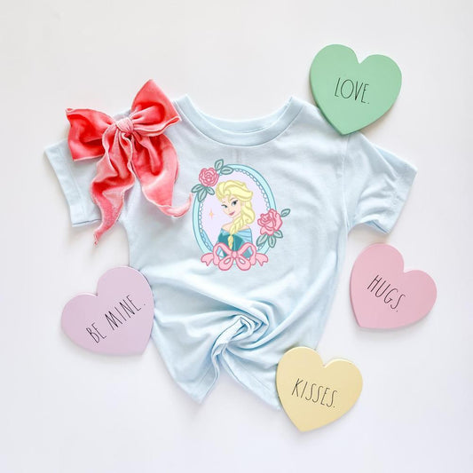 Light blue Snow Queen T-shirt with Elsa in a floral frame, a pink velvet bow, and heart-shaped "LOVE," "HUGS," "KISSES," and "BE MINE" candies.