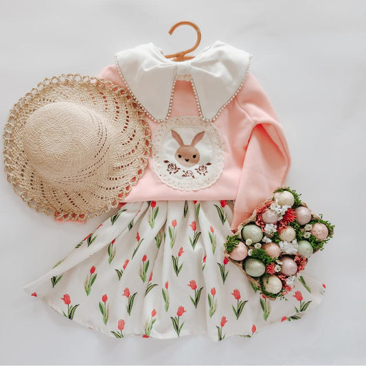 Pink knitted Snuggly Bunny Sweater with white collar, paired with a white tulip-print skirt, straw hat, and Easter eggs.
