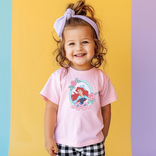Smiling child in a pink "Under the Sea" t-shirt featuring Ariel in a floral frame, with black and white plaid pants.