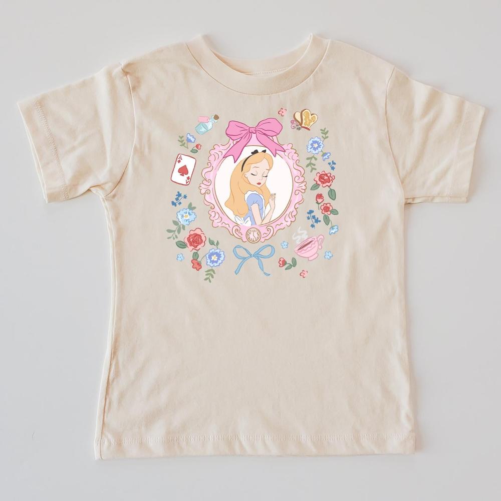 Beige Wonderland kids' t-shirt with Alice in a floral frame, pink bow, playing cards, and teacup design.