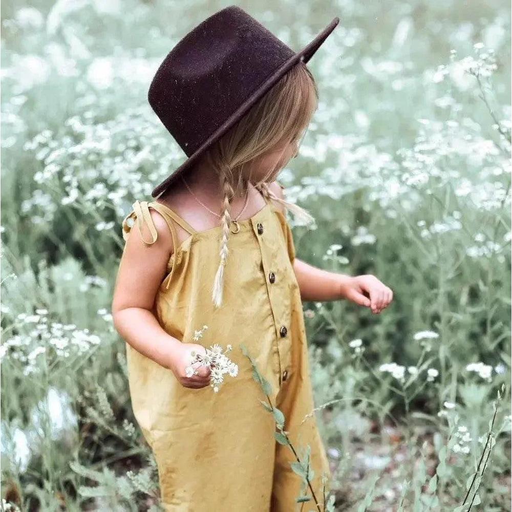 Toddler in mustard romper and wide-brim hat picking wildflowers in a grassy field