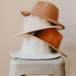 Stack of three classic brim hats in tan, cream, and brown on a stool, minimal background