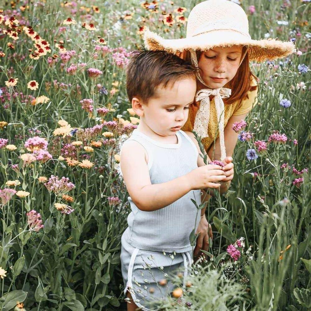 Young boy in light clothes and girl in straw hat exploring wildflowers in a sunlit meadow.
