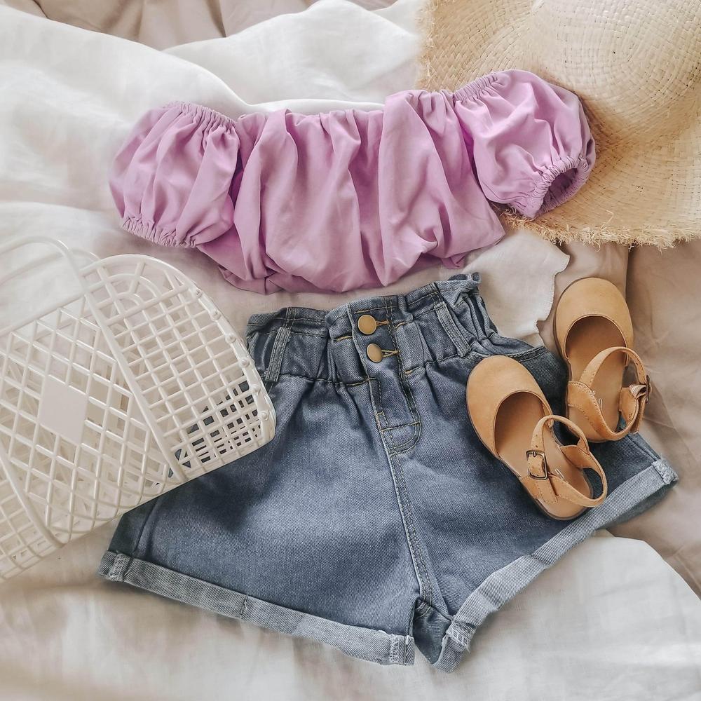 Girls’ summer outfit with a lilac puff sleeve top, denim shorts, tan sandals, sun hat, and white basket bag