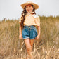 Smiling girl in straw hat, yellow blouse, and denim shorts standing in a grassy field.