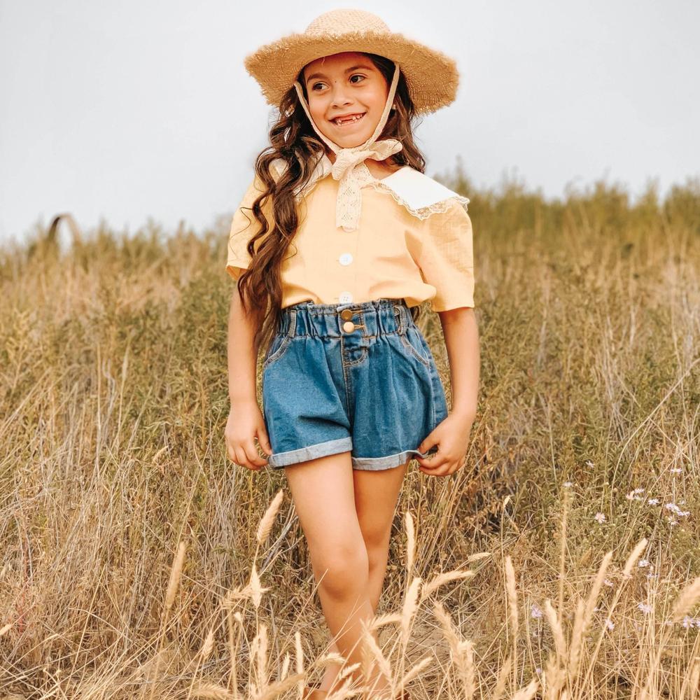 Smiling girl in straw hat, yellow blouse, and denim shorts standing in a grassy field.